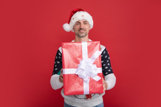 Portrait of santa man in sweater holding big red present box\
isolated over red background concept of christmas holidays\
happiness emotions facial expression joy and celebration