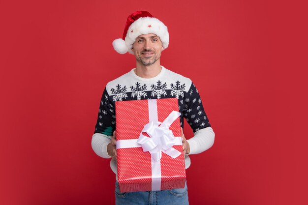 Portrait of Santa man in sweater holding big red present box isolated over red background Concept of Christmas holidays happiness emotions facial expression joy and celebration