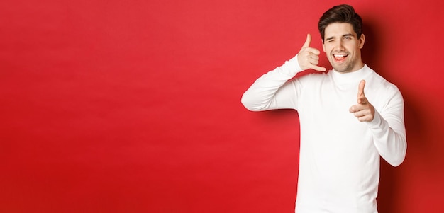 Portrait of sassy and confident handsome man flirting with you asking to call him showing phone sign and pointing at camera standing over red background