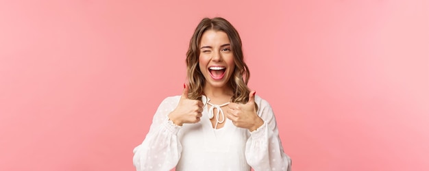 Portrait of sassy goodlooking blond girl assure all good show thumbsup and wink with classy pleased smile recommend visit place shop at online store standing in white dress pink background