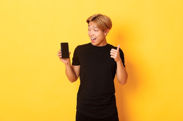 Portrait of satisfied asian guy looking at smartphone screen and showing thumbs-up in approval