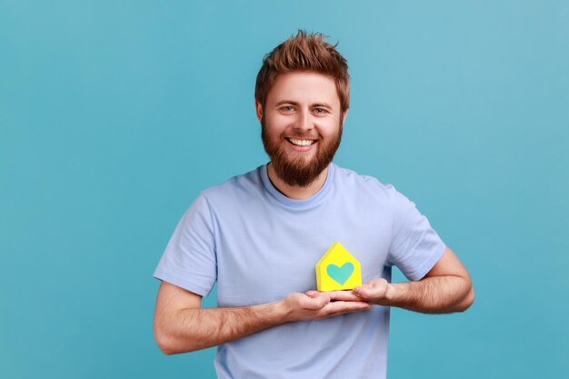 Portrait of satisfied bearded man holding in hands near heart little paper house looking at camera with pleasant smile, dreaming about own house. Indoor studio shot isolated on blue background.