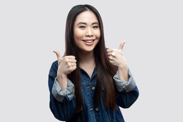 Portrait of satisfied beautiful brunette asian young woman in casual blue jacket with makeup standing, thumbs up, toothy smiling and looking at camera. studio shot, isolated on light grey background.