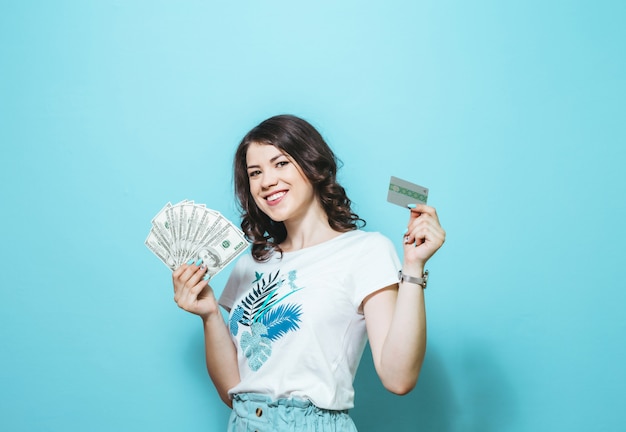 Portrait of a satisfied beautiful girl  holding money banknotes and credit card isolated over blue background