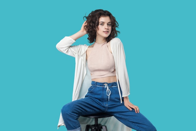 Portrait of satisfied beautiful young brunette woman in casual style sitting on chair and posing like fashion model and looking at camera with smile. indoor studio shot, isolated on blue background.