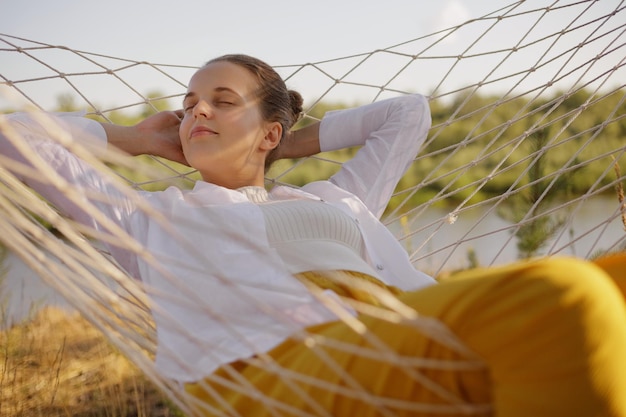 Portrait of satisfied caucasian young adult woman having nap in\
hammock on the bank of the river lying with closed eyes sleeping\
breathing fresh air wearing stylish clothing