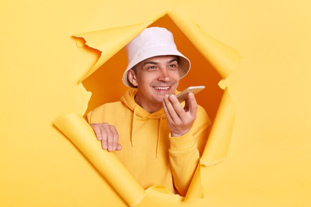 Portrait of satisfied delighted man wearing casual hoodie and panama looking through torn hole in yellow paper talking to cell phone using virtual assistant digital speaker application