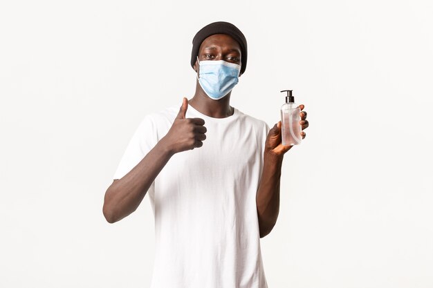 Portrait of satisfied handsome african-american man in beanie and medical mask, showing thumbs-up and recommend hand sanitizer, advertising