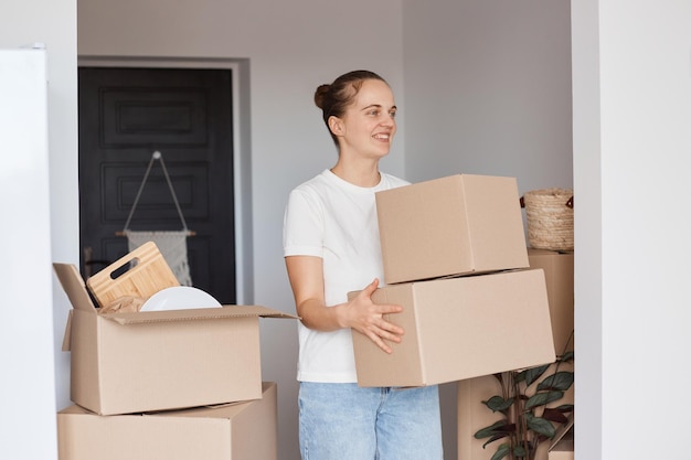 Portrait of satisfied happy beautiful woman wearing white casual style T-shirt and jeans standing with cardboard box with belongings, holding her personal stuff, looking away with smile.