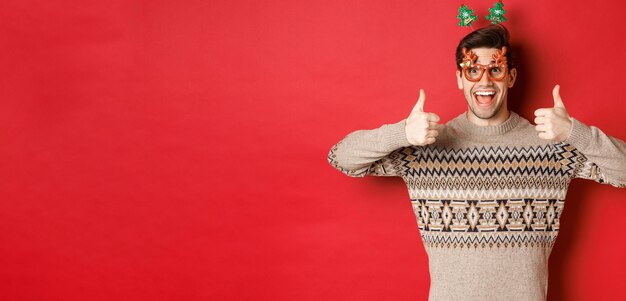 Portrait of satisfied and happy man in christmas sweater and party glasses, showing thumbs-up, wishing happy new year, standing over red background.