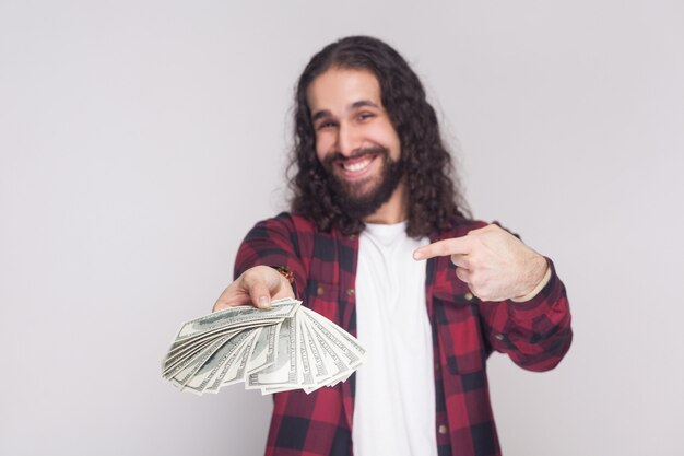 Portrait of satisfied happy young businessman in red checkered shirt and black long curly hair standing, giving and pointing with finger fan of money. Indoor studio shot, isolated on gray background