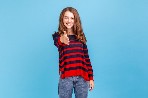 Portrait of satisfied pleased young woman wearing striped casual style sweater outstretching hand, offering handshake on job interview. Indoor studio shot isolated on blue background.