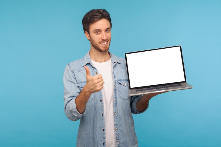 Photo portrait of satisfied smiling handsome man in worker denim shirt showing thumbs up and holding laptop screen with empty display, mock up for commercial image. studio shot isolated on blue background