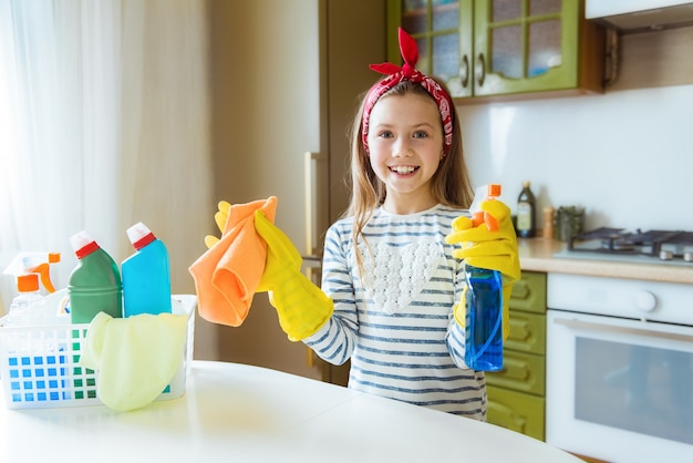 Photo portrait of satisfied teen girl housewife or cleaning lady working in the kitchen wearing an apron and gloves, holding a rag and detergent in her hands, looking at camera, smiling