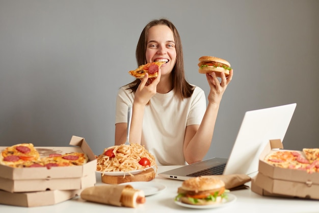 ラップトップ コンピューターとおいしいジャンク フード ディナーを楽しんでいるピザをかむ灰色の背景の上に分離された別の不健康な食べ物でテーブルに座って満足している女性の肖像画