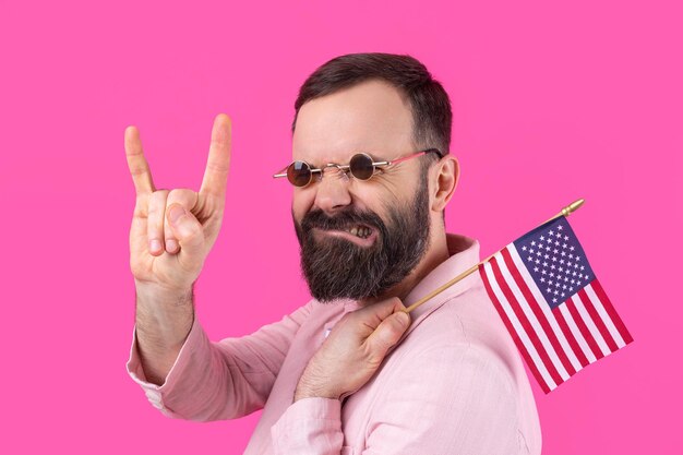 Portrait of a satisfied young man with a beard with an american\
flag on a red studio background great us patriot and defender of\
freedom