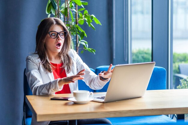 Ritratto di giovane donna bruna bella ed elegante spaventata con gli occhiali seduta guardando e leggendo alcune notizie sullo schermo del suo laptop con faccia impaurita scioccata in studio interno girato sfondo caffè ufficio