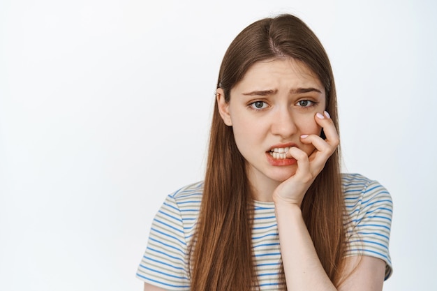 그녀의 손가락을 물고, 불안하고 걱정하는 카메라를 찾고, 흰색 티셔츠에 서있는 겁 먹은 소녀의 초상화. 복사 공간