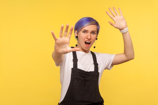 Portrait of scared hipster woman with violet short hair in denim overalls showing stop gesture and looking surprised frightened shouting from sudden fear isolated on yellow background studio shot