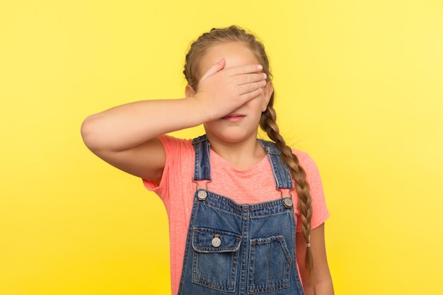 Портрет испуганной расстроенной маленькой девочки с косой в джинсовом комбинезоне, закрывающей глаза рукой, ребенок чувствует стыд или страх перед просмотром запрещенного контента в закрытой студии, снятой на желтом фоне