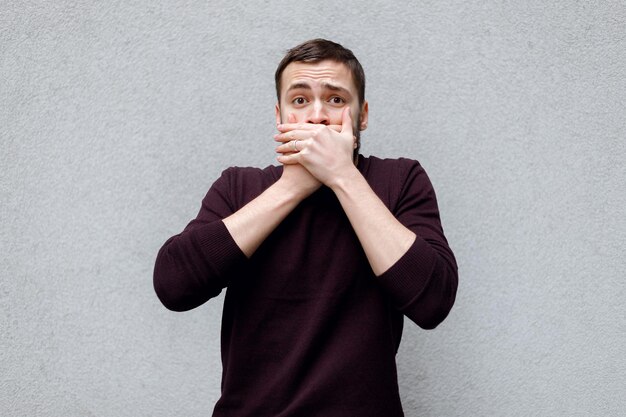 Portrait of scared young man covering with hand his mouth isolated on gray wall background. human emotions face expression