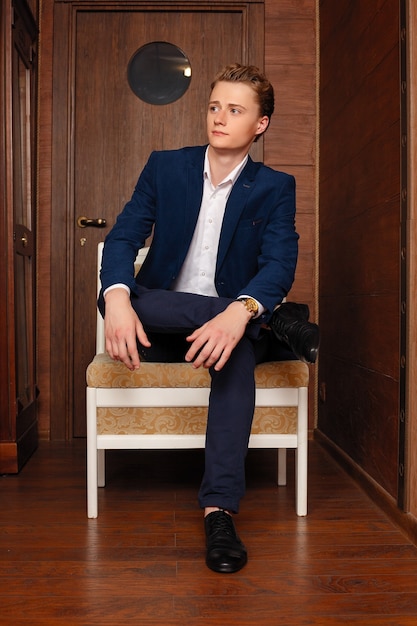 Portrait of School Boy. Dressing formally in black suit, necktie, white shirt, hands in pockets, a young handsome college student standing against vintage style office door on campus, looking at you.