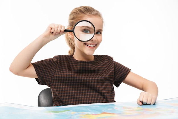 Foto ritratto di una ragazza della scuola che guarda la mappa del mondo attraverso la lente d'ingrandimento mentre studia geografia a scuola isolata su un muro bianco