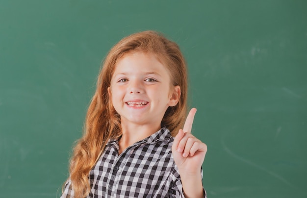 Ritratto dell'allievo nerd della ragazza della scuola con l'espressione sorprendente che indica con il dito contro la lavagna.