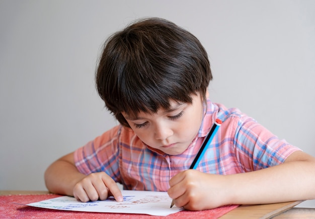 宿題をしているテーブルの上に立地する学校の子供男の子の肖像画、鉛筆書きを保持している幸せな子供、テーブル、小学校、ホームスクーリングのコンセプトで白い紙に描く少年