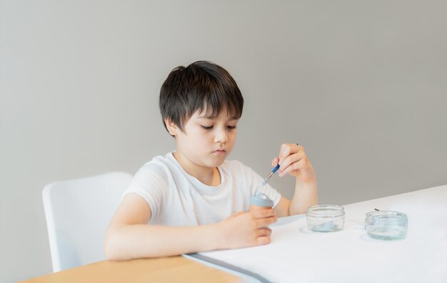 Photo portrait school kid using paint brushes painting grey colour on egg and diy easter decorations homework child boy preparing and getting ready for easter hunt for celebration on school holiday