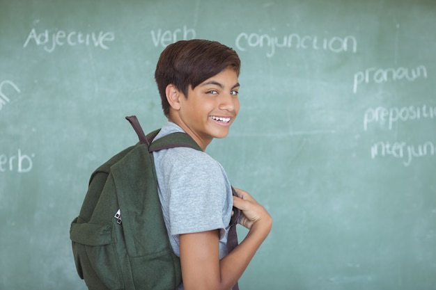 Portrait of schoolboy with backpack standing against chalkboard in classroom