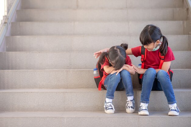 Portrait of schoolgirl crying while sitting on stairs outdoors with smiling sister comforting her, copy space