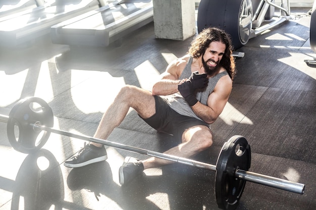 Portrait of screaming young adult man athlete with long curly hair working out in gym, sitting on floor and have strong hurt problem with wrist, spasm painful. Compressing with hand, screaming. Indoor