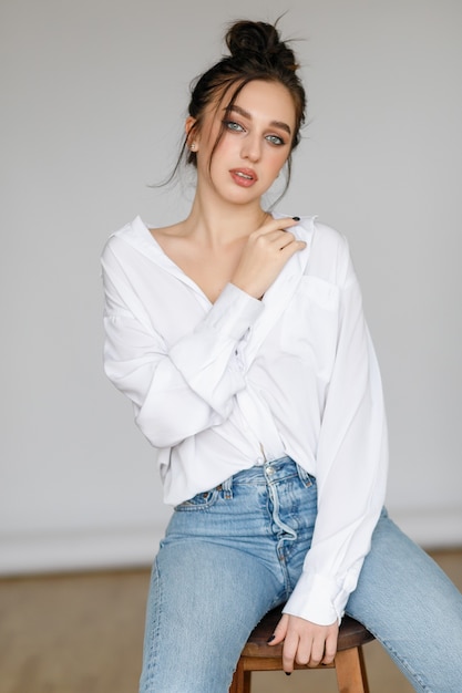 Portrait of seated young woman wearing a bouffant shirt with naked shoulders on light grey background. Beautiful brunette girl in jeans and white t-shirt smiling