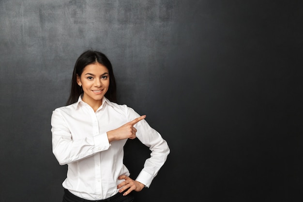 Portrait of secretary or business woman wearing white shirt pointing finger aside on copy space, isolated over dark gray wall