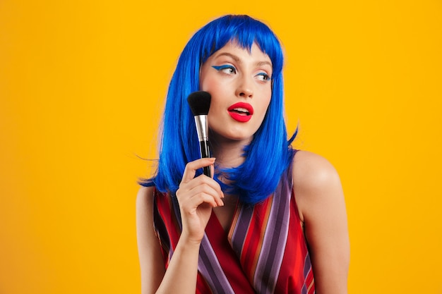 Portrait of seductive beautiful woman wearing blue wig looking aside and applying powder with brush isolated over yellow wall