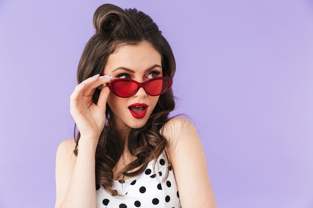 Portrait of seductive pin-up woman in retro sunglasses smiling and posing at camera isolated over violet wall