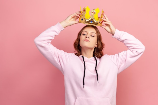 Portrait of selfish curly haired teenage girl in hoodie holding gold crown over head looking with arrogance privileged status Indoor studio shot isolated on pink background