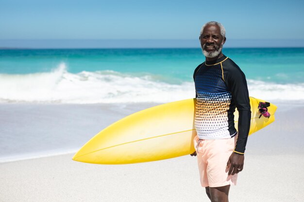 Portrait of a senior African American man on a beach in the sun, carrying a surfboard under his arm, looking to camera and smiling, with blue sky and sea in the background