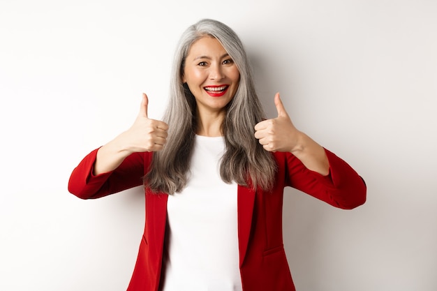 Portrait of senior asian woman showing thumb-up in approval, wearing red blazer for office work, recommend company, standing over white background