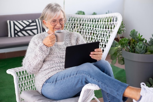 발코니에 있는 안락의자에서 휴식을 취하고 있는 디지털 태블릿을 사용하는 매력적인 노부인의 초상화