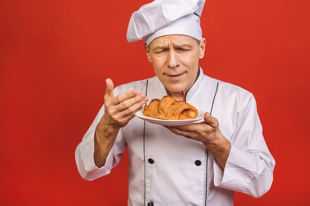 A portrait of a senior baker holding croissant isolated on red background.