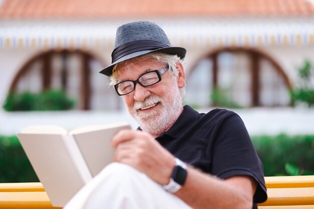 Portrait of a senior bearded man with hat sitting on a bench in the park relaxing and reading a book