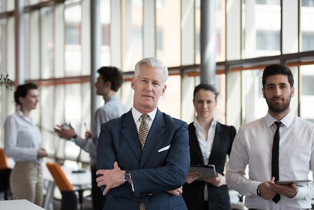 portrait of senior businessman as leader  at modern bright office interior, young  people group in background as team