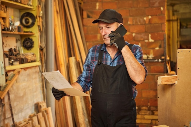 Portrait of a senior carpenter looking at blueprints plans to make a piece of furniture and talking on the phone with a client in the carpentry workshop