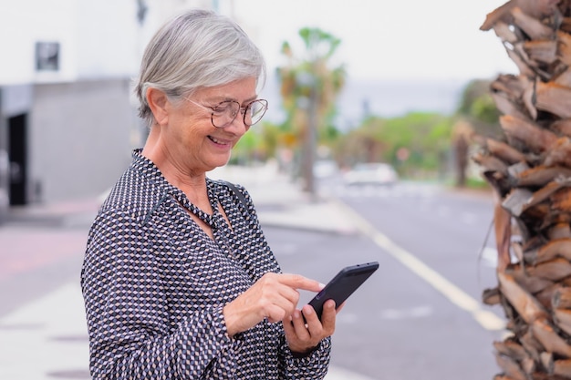 Portrait of senior confident caucasian businesswoman standing outdoor with city in background using mobile phone