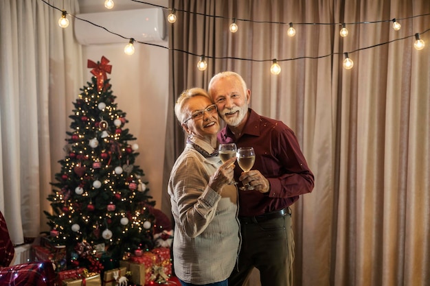 Портрет пожилой пары, тостующей с вином дома возле рождественской елки на Рождество