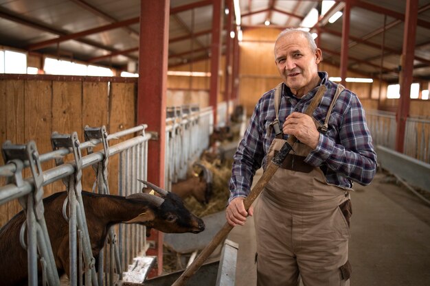 Photo portrait of senior farmer with pitchfork standing in farmhouse taking care and feeding domestic animals