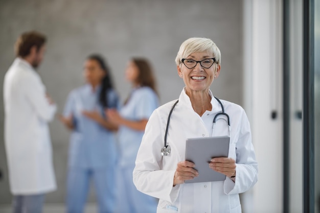 Portrait of a senior female doctor is looking at camera while working on a digital tablet in a hospital hallway.