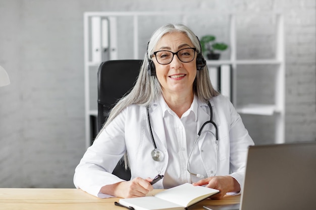 Portrait of senior grey-haired female doctor in her office using laptop for video chat with a patient. Online consultation with doctor for diagnoses and treatment recommendation. Telehealth concept.